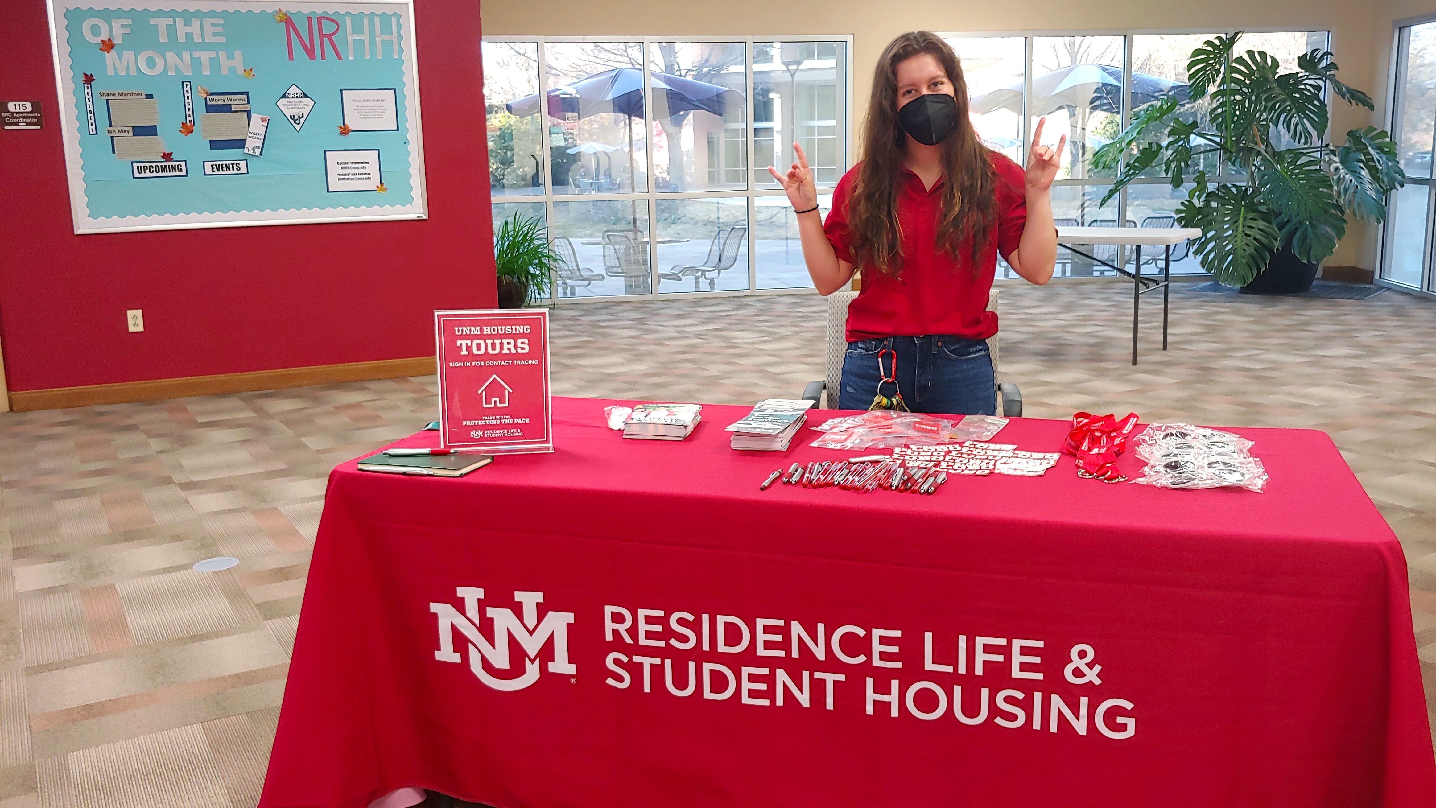Female standing behind a Residence Life and Student Housing table with her arms up giving the Lobos sign