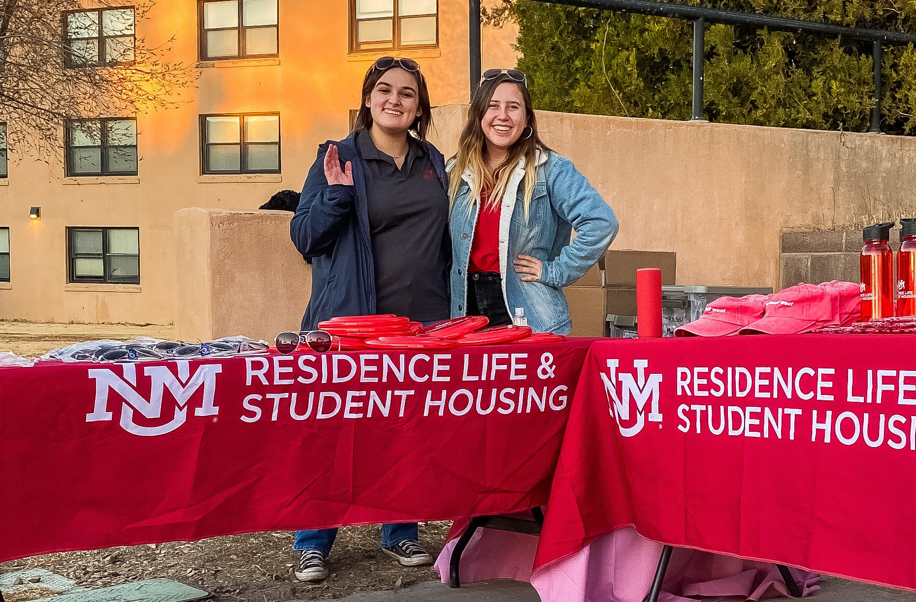 Two females smiling and waving standing behind a table with UNM Residence Like and Studetn Housing banner over it.
